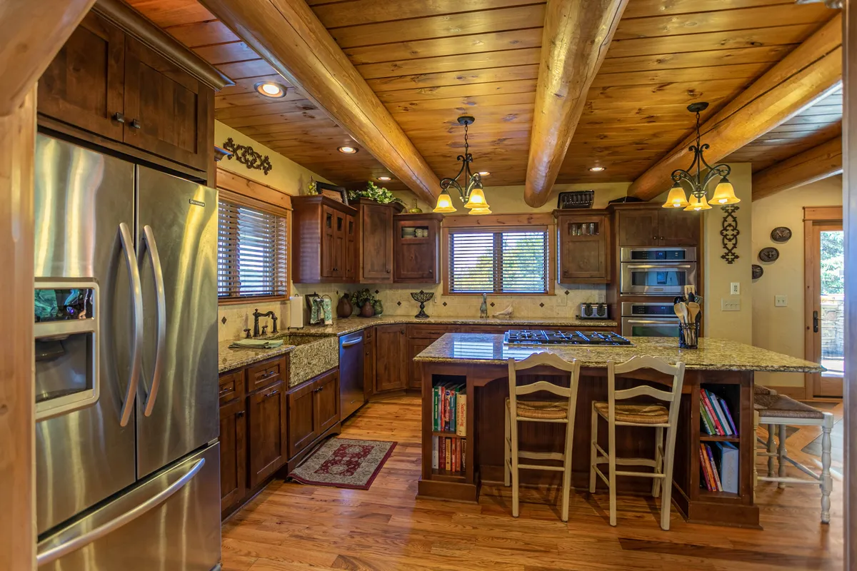 Featuring an open-concept kitchen with a central island, bar stools, stainless steel refrigerator, wooden cabinetry, granite countertops, and pendant lighting, this image radiates a cozy and communal feel ideal for entertaining and meal preparation.