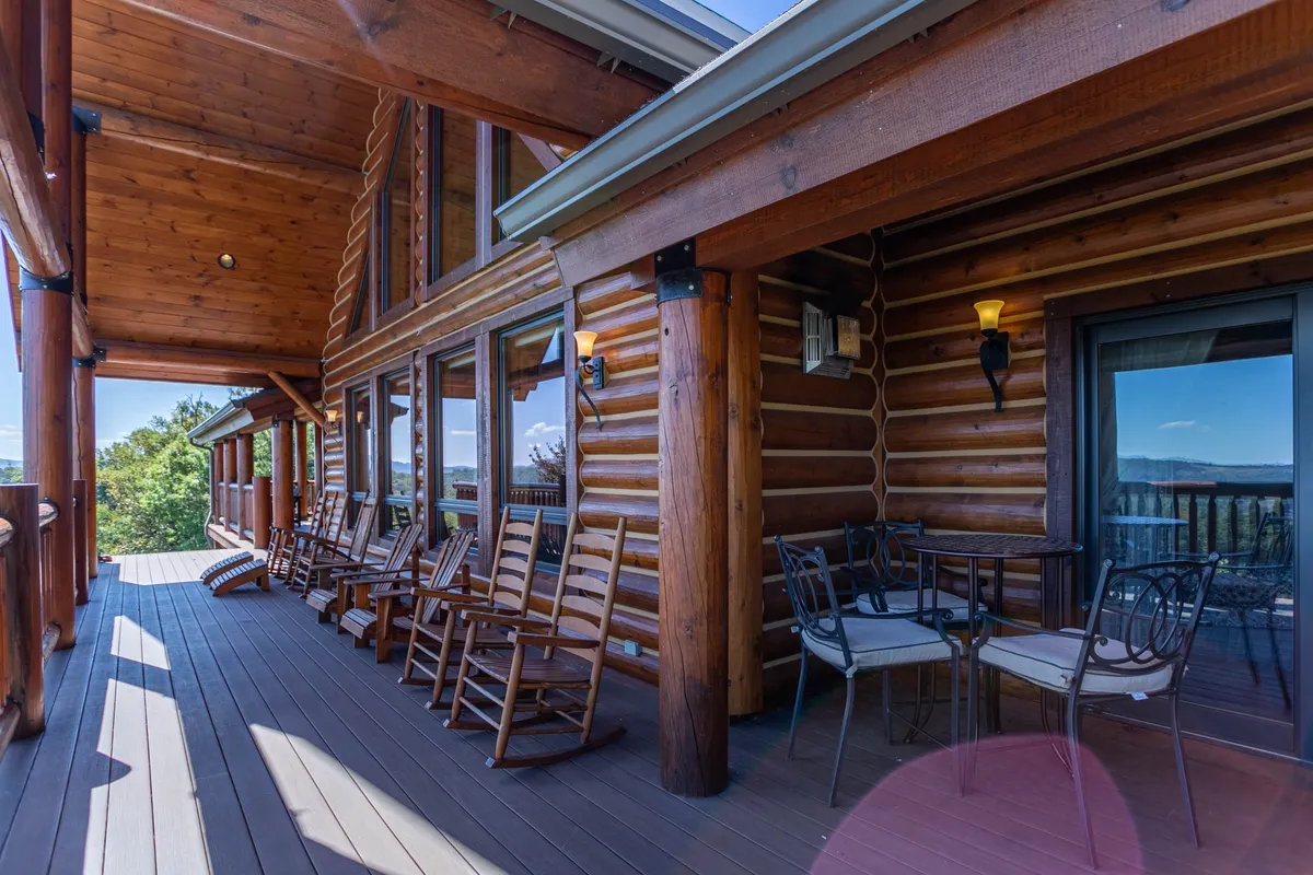 A tranquil exterior scene showing a wrap-around deck with wooden beams, rocking chairs, an outdoor table, and expansive panoramic views of the surrounding mountains and forest. The setting is ideal for morning coffee, alfresco meals, or sunset watching.