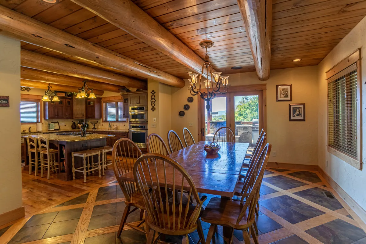 This image presents a large wooden dining table beneath rustic beams, a striking chandelier, and a view into the adjacent kitchen area. The setting is tailored for family dinners, celebrations, and socializing in a warm, communal space.