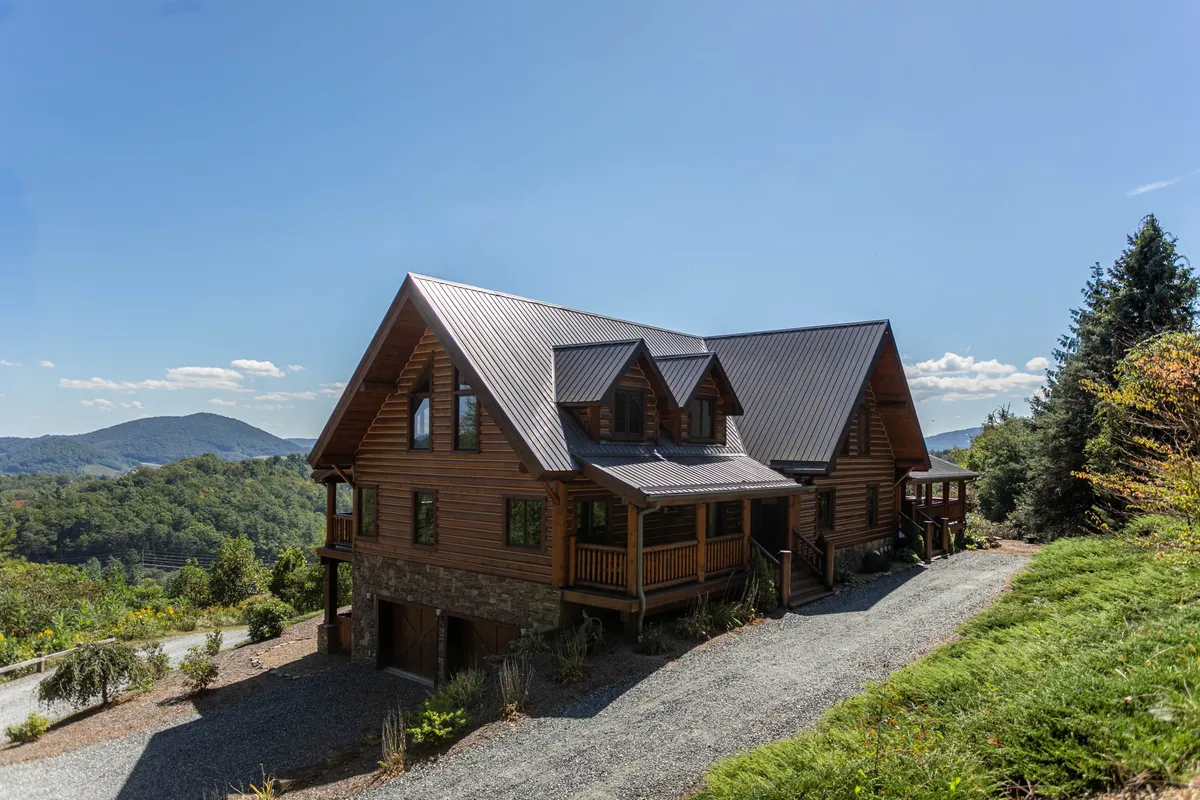 The hero image features a stunning log cabin set against a dramatic mountain backdrop. The surrounding green landscaping creates a peaceful and inviting atmosphere, while the spacious gravel driveway hints at ample room for guests. An expansive porch offers a place to relax and take in the breathtaking sunset views, evoking feelings of serenity and togetherness.