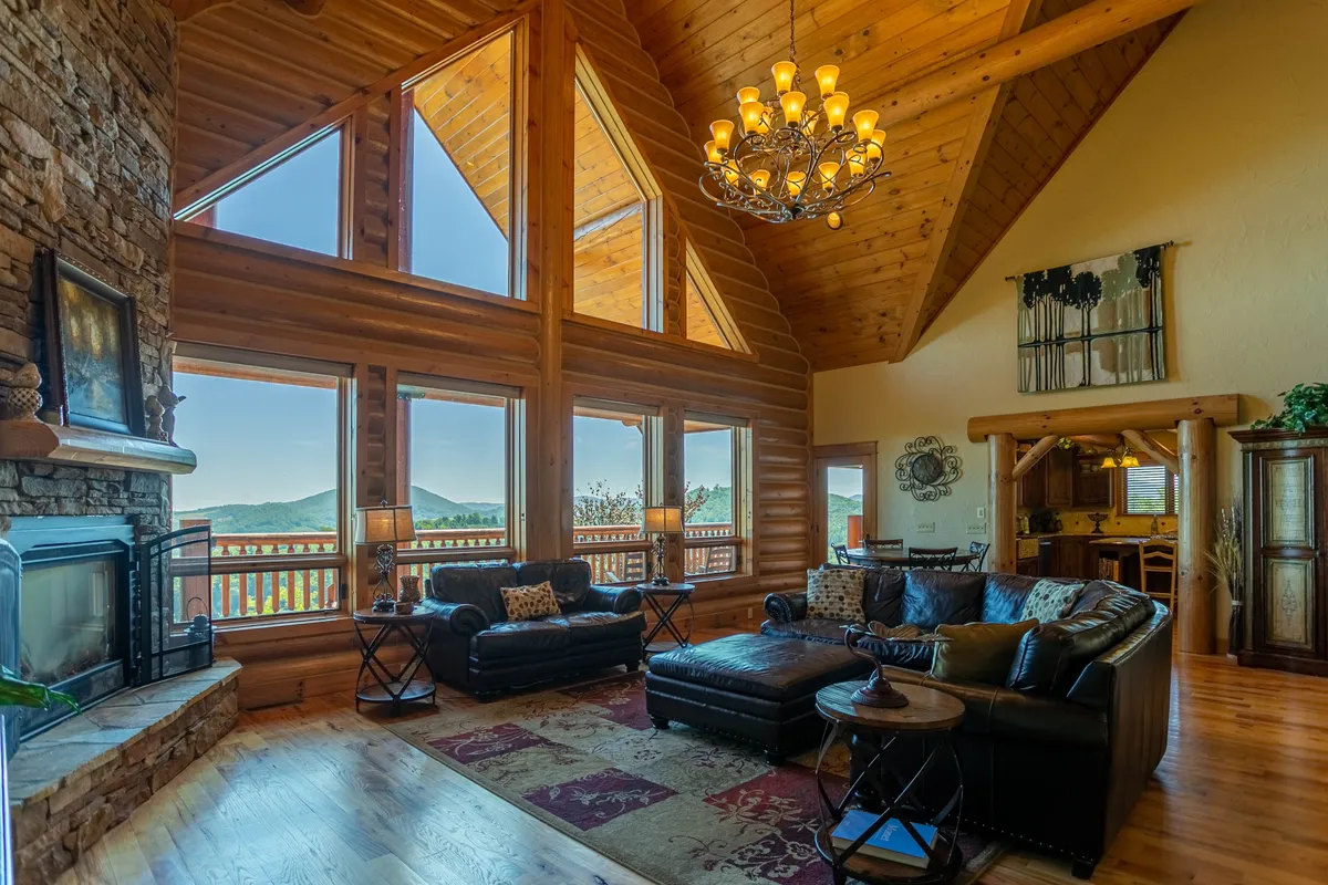 A spacious great room with soaring ceilings, a grand chandelier, large windows framing panoramic mountain views, a central stone fireplace, and plush leather sofas arranged for relaxation and gathering. Sunlight pours in, creating a warm, cozy atmosphere perfect for enjoying the landscape in any season.