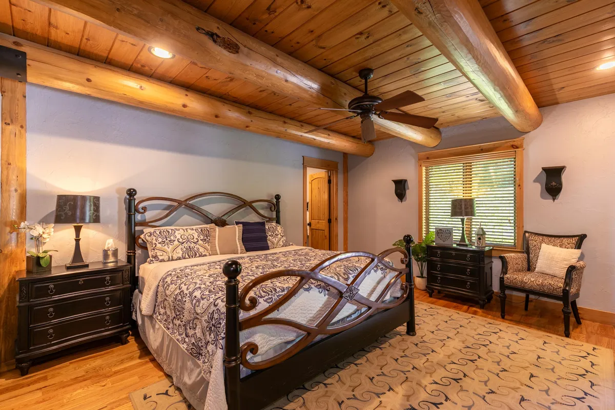 A bedroom featuring a bed with a decorative headboard, cozy nightstands with lamps, exposed wooden beams, and a window allowing sunlight to stream in. The setting is warm and relaxing, perfect for deep rest or unwinding with a book.