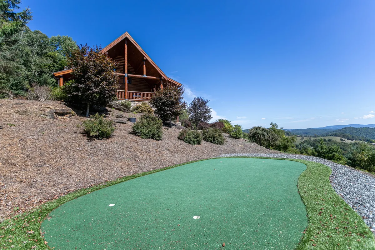 An inviting log cabin exterior with a lush putting green in the foreground and expansive mountain views beyond. The golden light of sunset bathes the scene, evoking a sense of peace and fun outdoor activity. The putting green hints at friendly family competition and relaxation after a day of exploring local attractions.