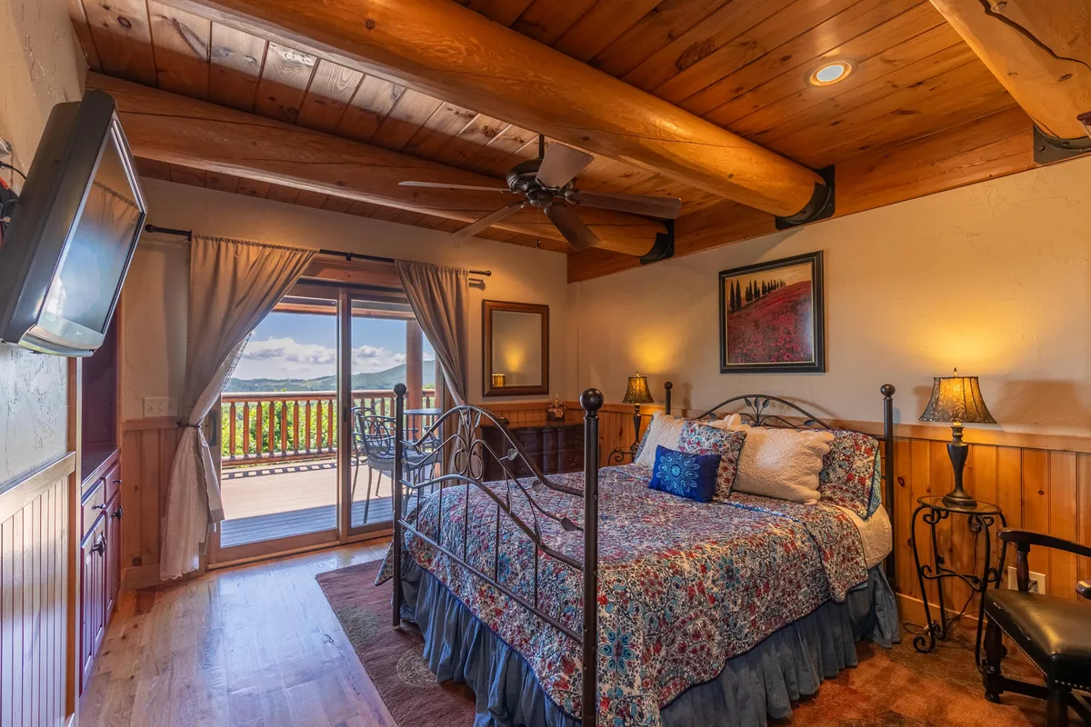 A serene bedroom featuring a bed with floral bedding, wooden beams overhead, and a large sliding door opening to the wrap-around deck. The scene is bathed in natural light, inviting guests to wake up to the sunrise or stargaze from the comfort of their room.