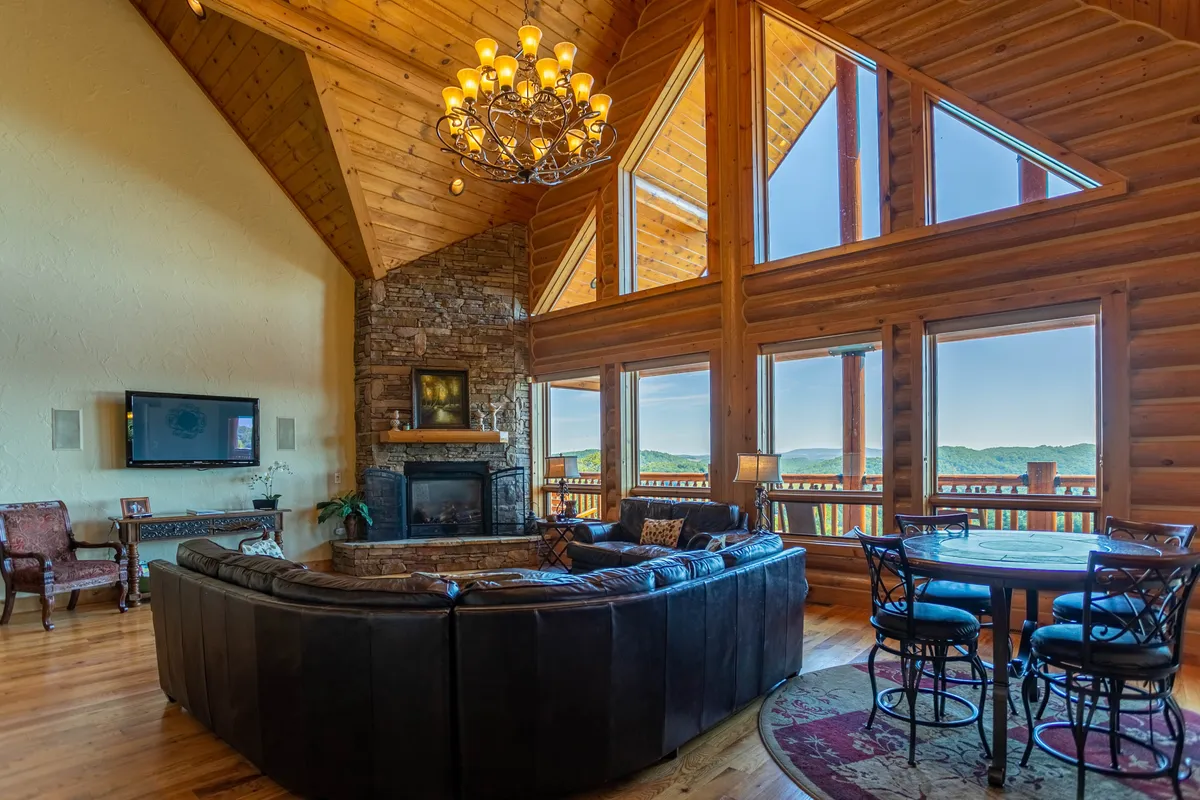 This image showcases a spacious great room with a prominent stone fireplace, expansive windows that frame breathtaking mountain views, a dramatic chandelier, a curved leather sofa, and a dining area. The atmosphere feels inviting and warm, emphasizing both the grandeur and comfort of the space.